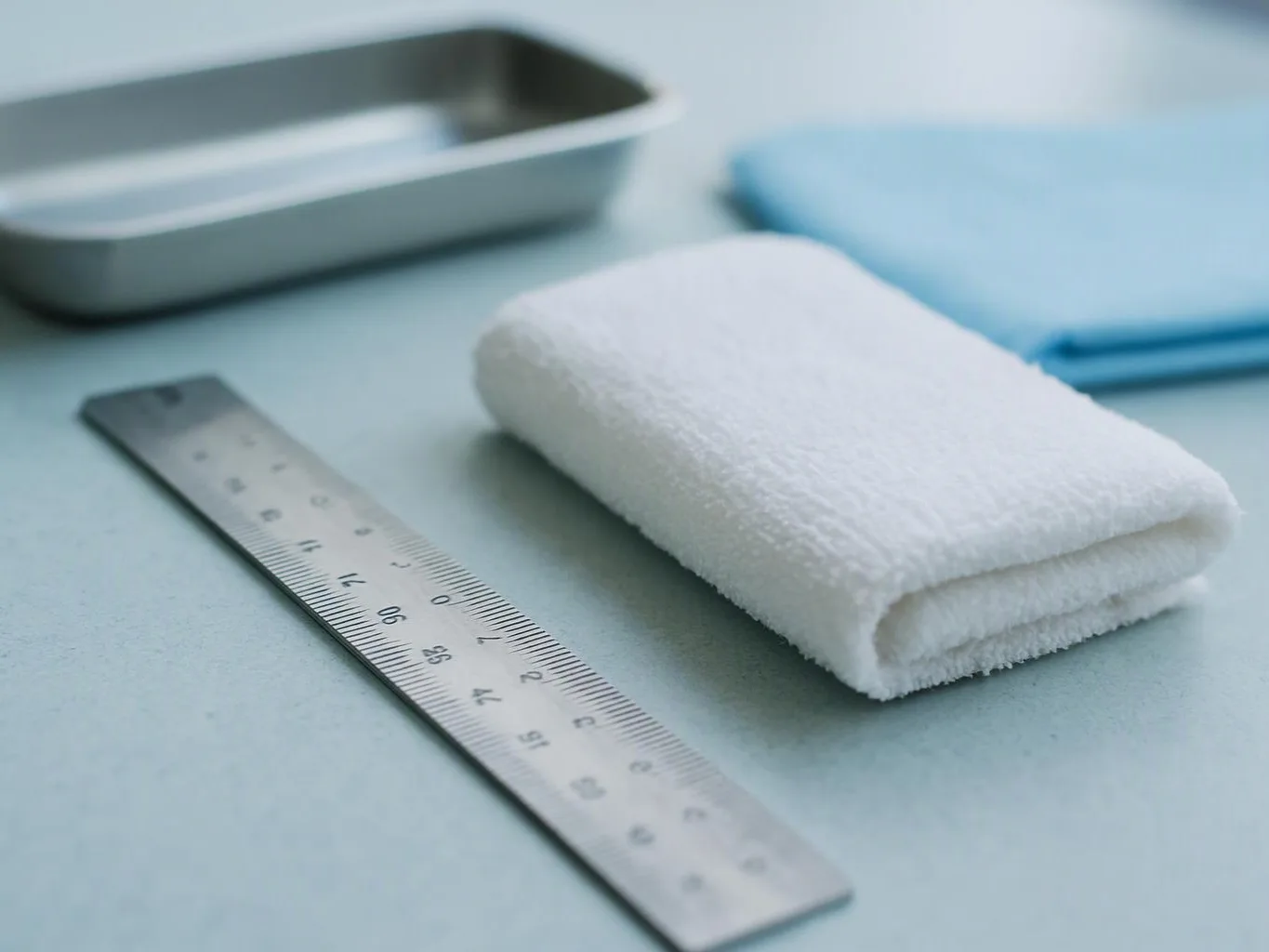 A folded white towel sits beside a metal ruler and tray on a pale blue surface