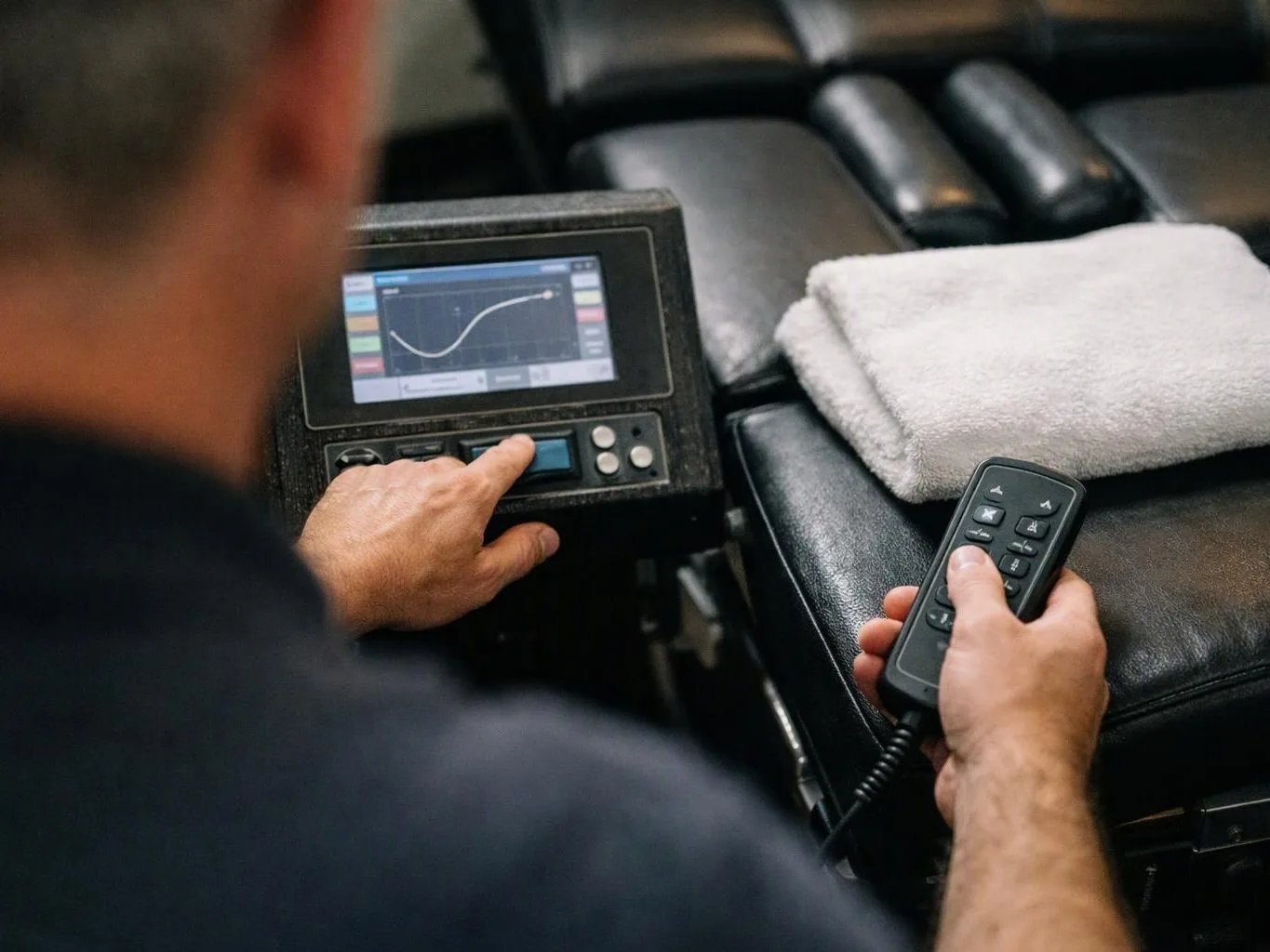 A person adjusts a massage chair controller while holding a remote, with a folded towel on the black cushion