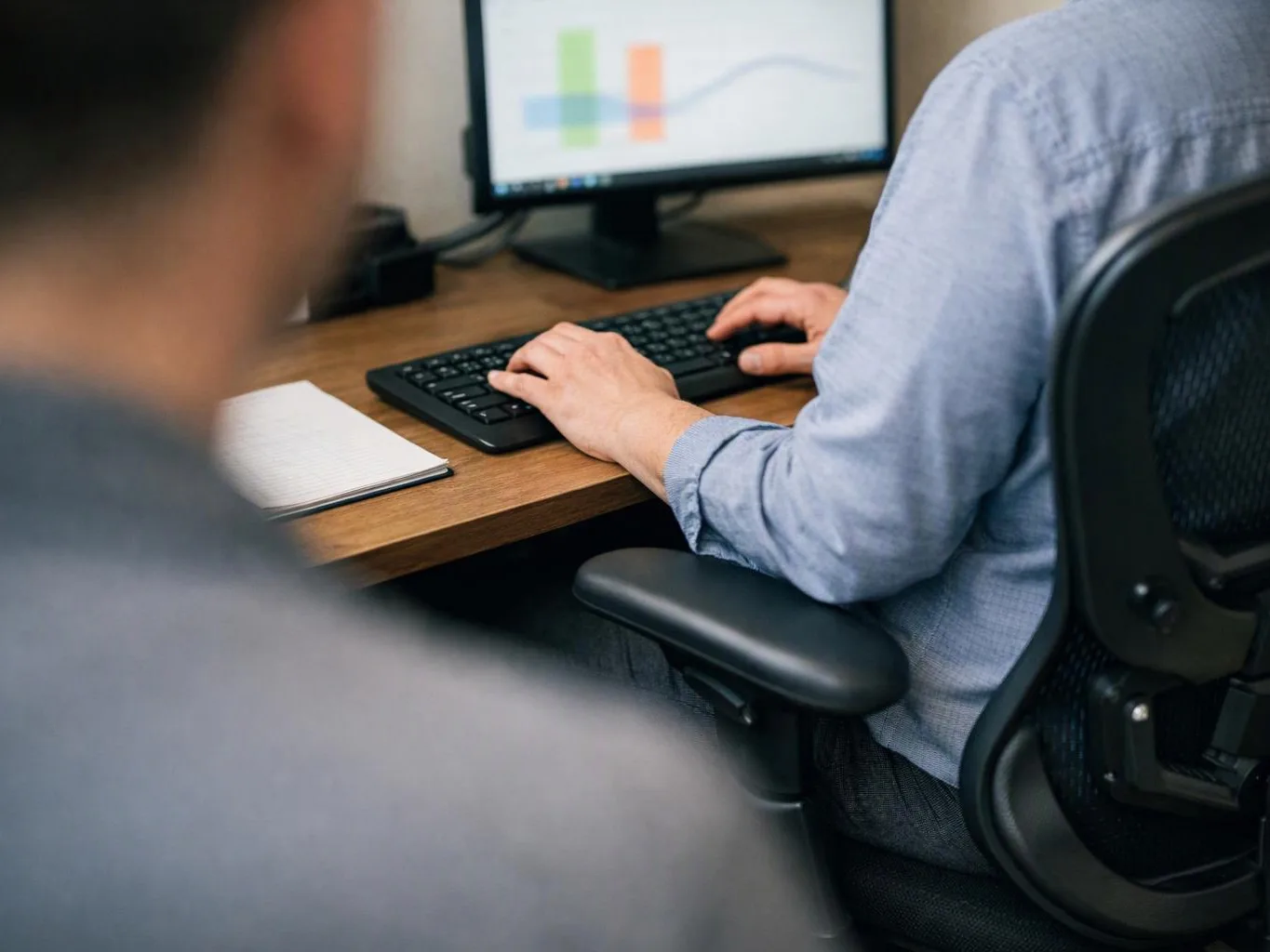 Two people sit at a desk, one typing on a keyboard while a monitor shows charts in an office setting